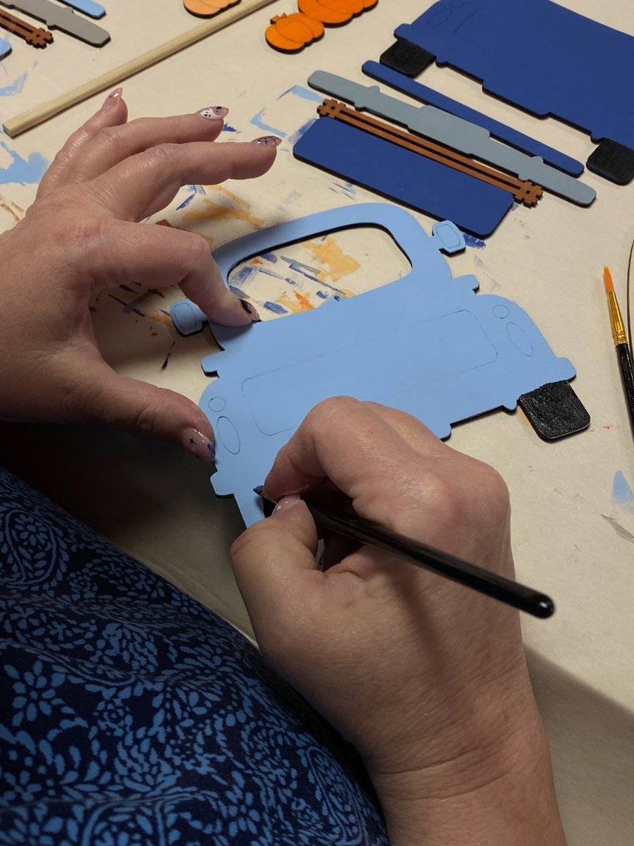 Hands painting a laser-cut blue wood truck kit at a Cricket Creek Studio workshop, with paint brushes and colorful wood pieces on the table